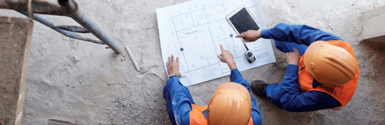 Construction managers in orange hardhats pointing at blueprint on the floor